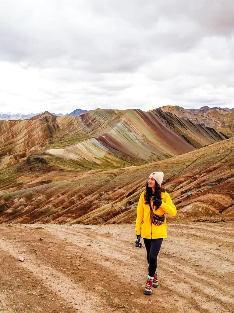 A female hiker in yellow jacket in front of rainbow coloured mountains