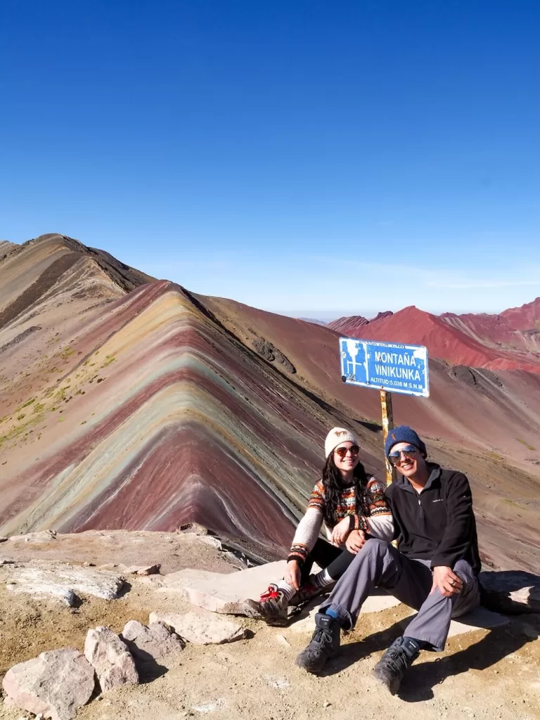 A hiking couple is sitting in front of the Vinicuna sign with a rainbow coloured mountain behind them