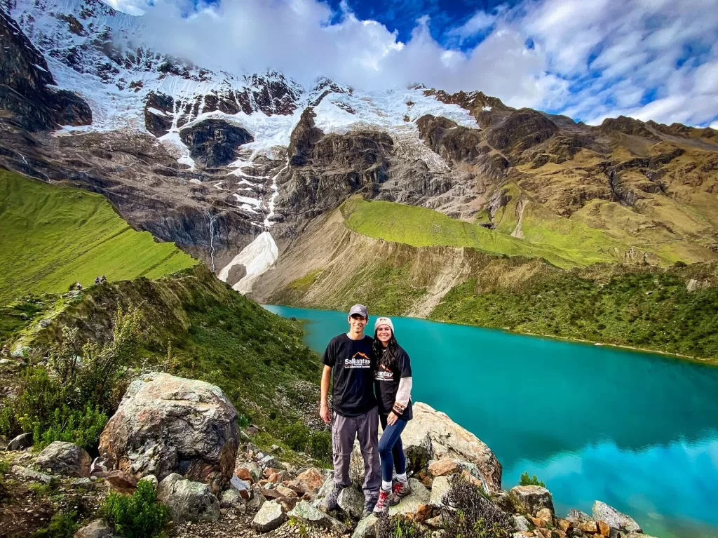 A male and female hiker couple standing on top of a ridge in front of an emerald lagoon and snowcapped mountain
