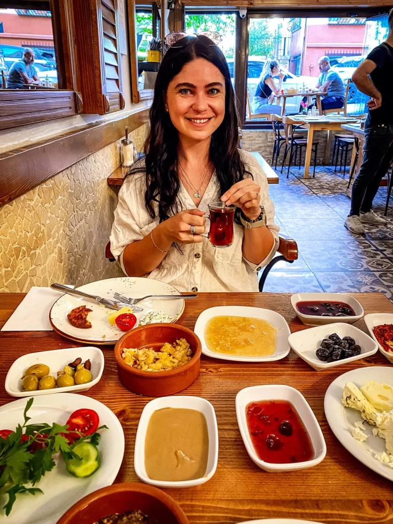 A smiling girl holding a glass of Turkish tea with a selection of food in front of her on a table