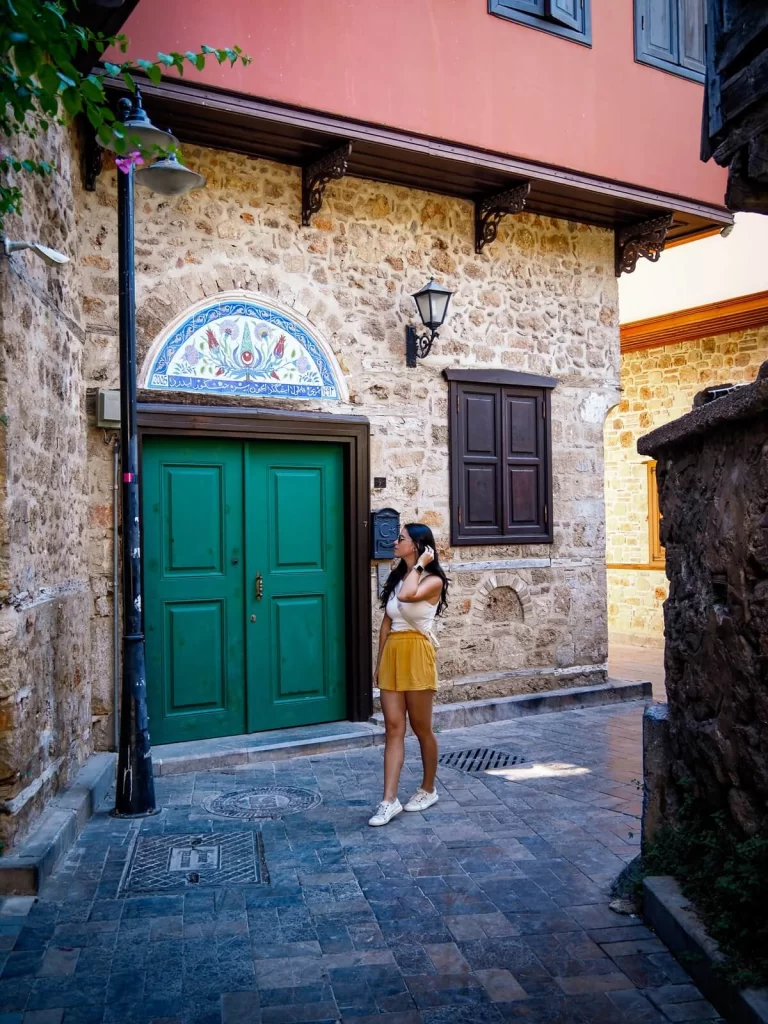 A female traveller wearing yellow shorts and a white shirt is walking through a narrow cobblestone street in front of a greed double door