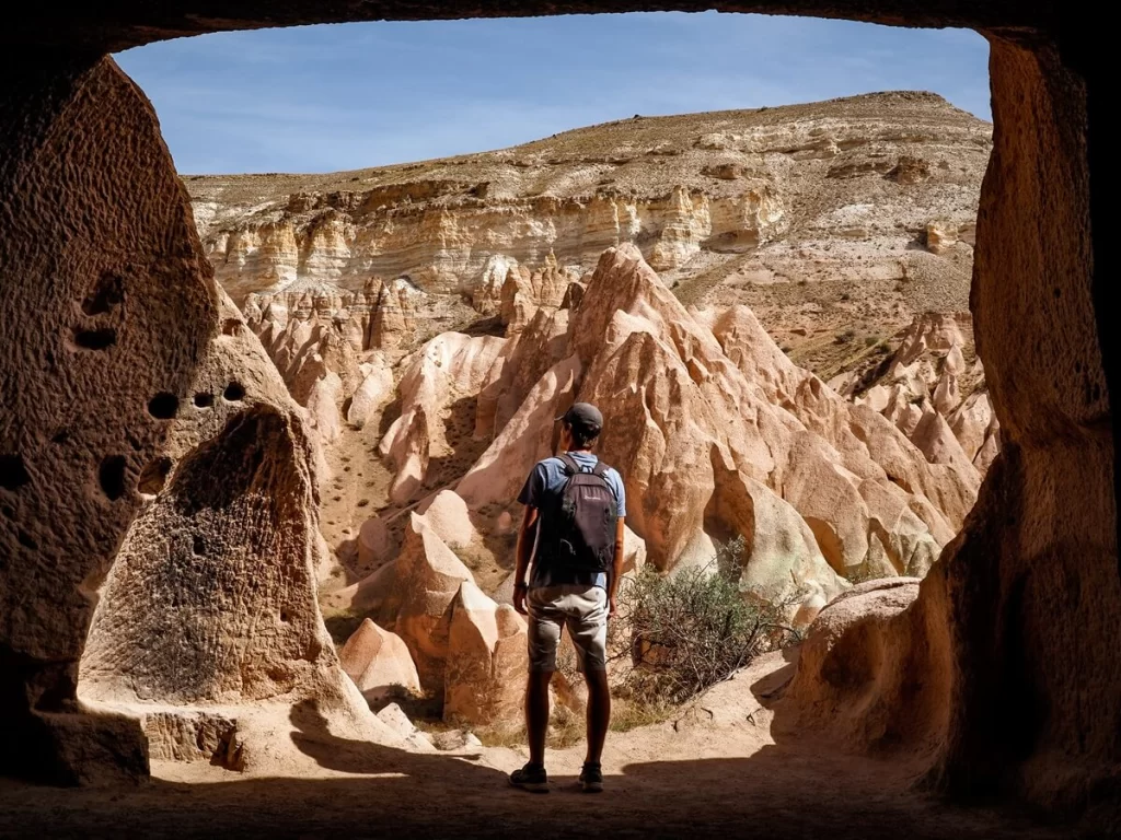 A male traveller is standing in a cave overlooking a valley with unique rock formations