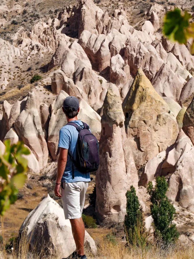 A male hiker wearing a backpack is standing in front of pink rock formations in Cappadocia Turkey