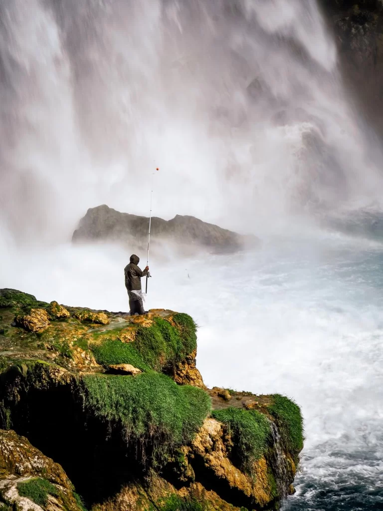 A fisherman fishing on mossy rocks in front of a waterfall