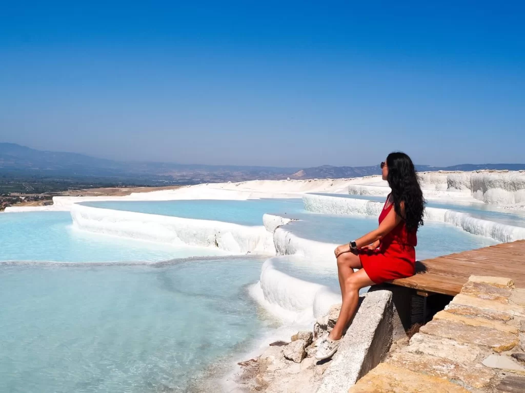 A female traveller in red dress sitting in front of white travertine pools filled with blue water in Pamukkale Turkey