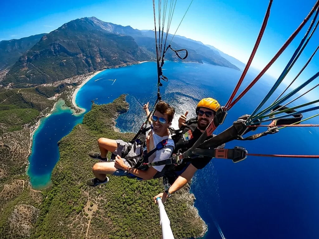 Male traveller and male instructor paragliding above a deep blue lagoon and the sea with lush green coast