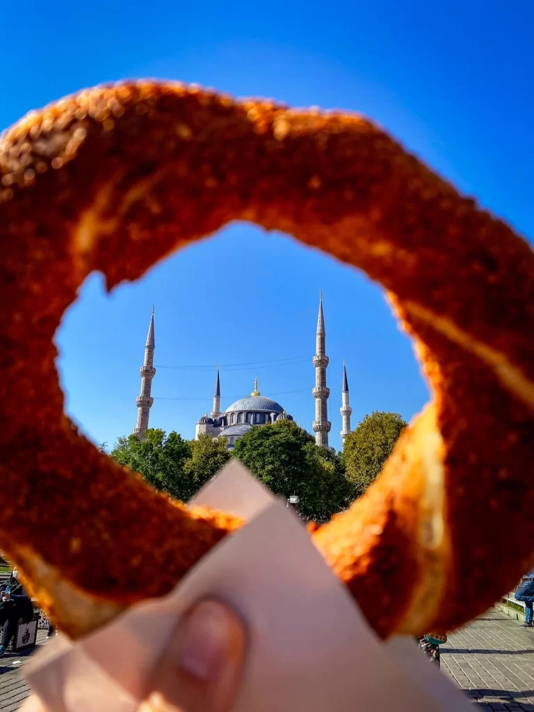 A hand holding up a bagel-like bread called simit with a mosque in the background