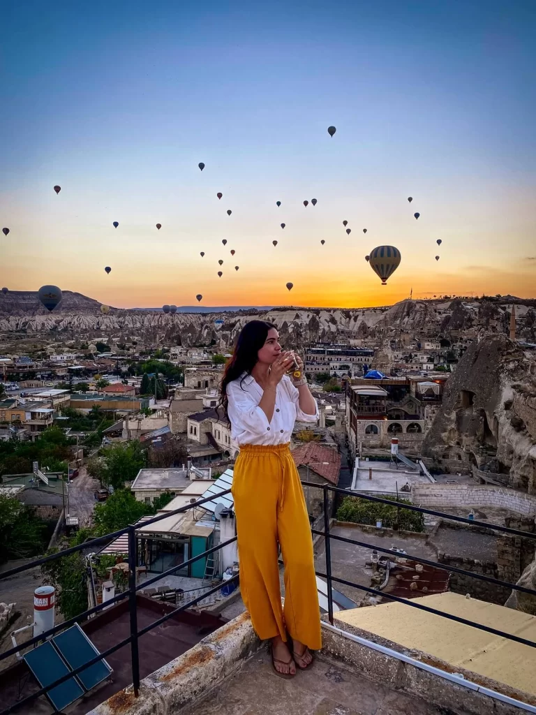 A woman in yellow trousers drinking tea on a balcony with hot air balloons in the background in Cappadocia Turkey