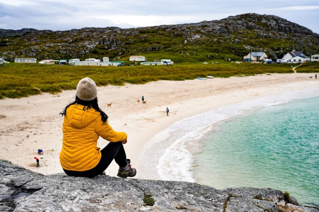 Female in yellow jacket is sitting on rocks overlooking Achmelvich Bay in Scotland