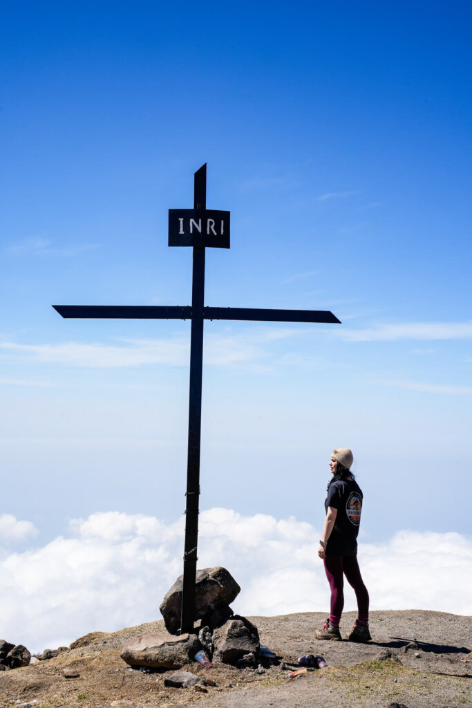 Female hiker is standing next to a cross on top of Santa Maria Volcano above the clouds