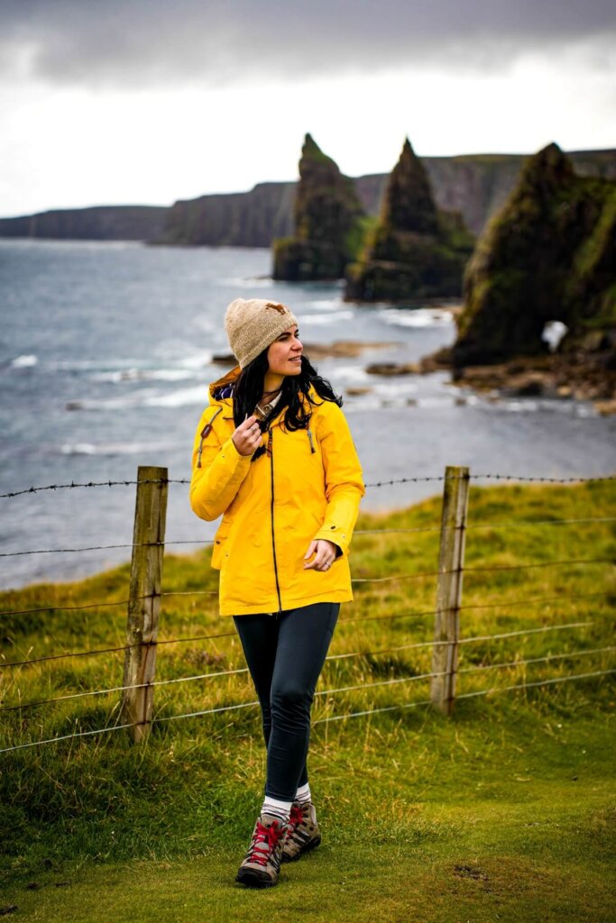 Female hiker in yellow jacket is standing in front of some sea stacks in Scotland