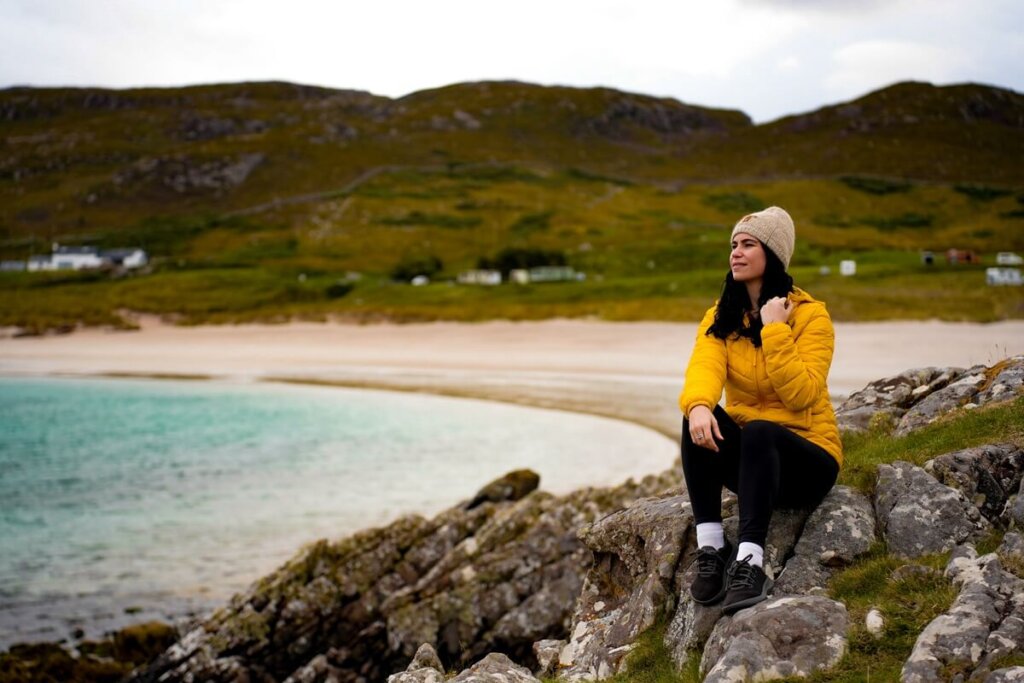 Female in yellow jacket is sitting on rocks at Mellon Udrigle Beach in Scotland