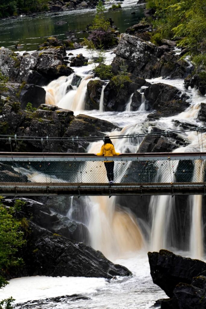 A female is standing on a hanging bridge in front of Rogie Falls in Scotland