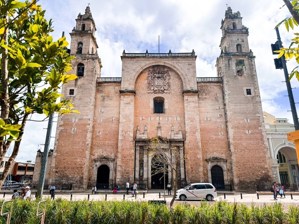 Front view of the Cathedral of Mérida in Yucatán, Mexico, showing its colonial facade with two bell towers, a central arch featuring a carved crest, and stone walls. A few people walk in front of the cathedral, and a white car is parked nearby, with green foliage and a partly cloudy sky in the background